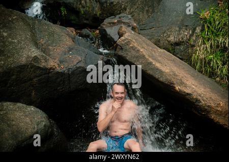 Ein Mann genießt eine natürliche Wasserfallmassage und lacht, während das Wasser in einer ruhigen Langkawi-Umgebung über ihn kaskadiert Stockfoto