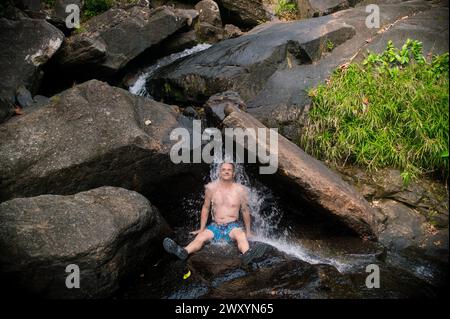 Ein Mann sitzt unter einem erfrischenden Wasserfall inmitten der zerklüfteten Felsen eines Langkawi-Dschungels, umgeben von üppigem Grün Stockfoto