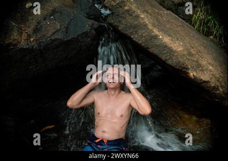 Ein Mann lacht mit purer Freude, während er eine natürliche Wasserfalldusche im Herzen von Langkawis üppigem Wald genießt Stockfoto