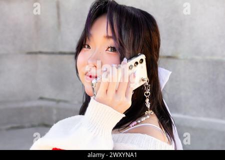 Ein modernes chinesisches Mädchen der Generation-Z posiert mit ihrem Smartphone und zeigt trendige Mode und Accessoires vor einem urbanen Hintergrund Stockfoto