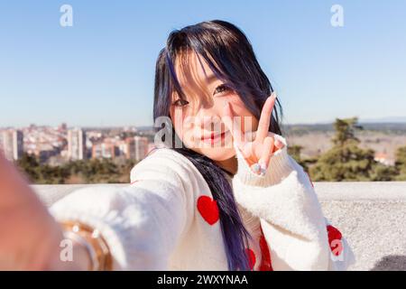 Ein chinesisches Mädchen mit gefärbten blauen Haaren und einem weißen Pullover mit roten Herzen macht ein Friedenszeichen, während es ein Selfie macht, mit einer Stadtlandschaft im Hintergrund Stockfoto