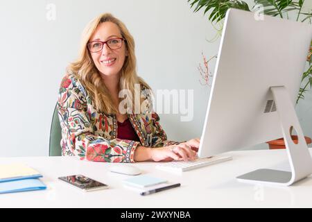 Eine professionelle blonde Geschäftsfrau mit einer Brille, die in die Kamera lächelt, während sie an einem Computer in einer modernen Büroumgebung arbeitet Stockfoto