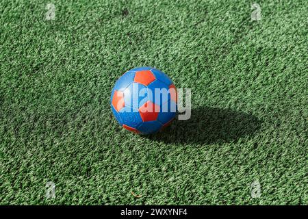 Ein leuchtend blau-roter Fußballball sitzt auf einem Bett aus künstlichem grünem Gras und vermittelt ein Gefühl von Spiel und Sport Stockfoto