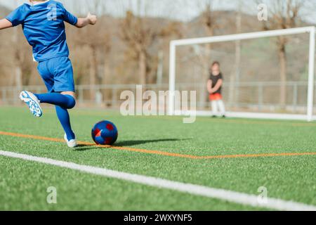 Ein junger Fußballspieler in Blau ist dabei, einen rot-blauen Ball auf einem leuchtenden grünen Feld zu treten, mit einem Torwart im Hintergrund Stockfoto