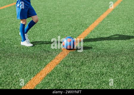 Zugeschnittene Ansicht eines Fußballspielers in Blau, der neben einem bunten Fußball auf dem grünen Kunstrasen steht, Schatten sichtbar Stockfoto