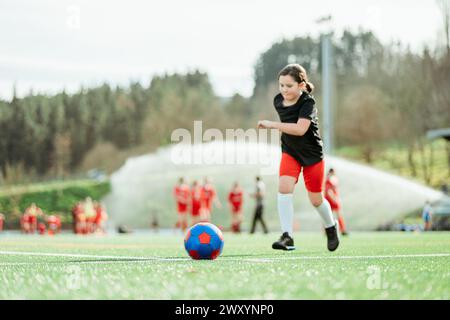 Ein junges Mädchen dribbelt selbstbewusst einen Fußball auf einem grünen Feld an einem sonnigen Tag, mit einem Team im Hintergrund Stockfoto