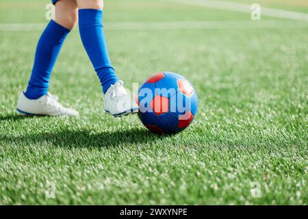 Die Beine eines Fußballspielers in blauen Socken und weißen Schuhen, die gerade einen rot-blauen Fußball auf einem grünen Feld treten Stockfoto