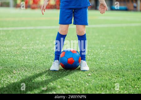 Eine Nahaufnahme eines Kindes in blauer Sportbekleidung, das auf einem grünen Fußballfeld steht, mit einem leuchtend rot-blauen Ball zwischen den Füßen Stockfoto