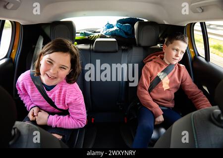 Zwei Kinder, ein lächelndes Mädchen und ein Junge, der aus dem Fenster blickt, werden während eines Familienausflugs auf dem Rücksitz festgeschleppt, und das Gepäck ist im Inneren angesammelt Stockfoto