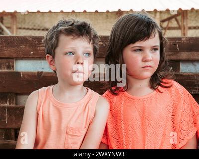 Ein Junge und ein Mädchen sitzen Seite an Seite an Seite an einem Holzzaun, verloren in Gedanken und genießen die Sehenswürdigkeiten einer Bauernschule während des Sommers Stockfoto