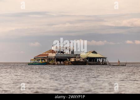 Rustikale schwimmende Struktur auf einem ruhigen Meer mit einem traditionellen Boot in der Nähe unter einem bewölkten Himmel. Stockfoto