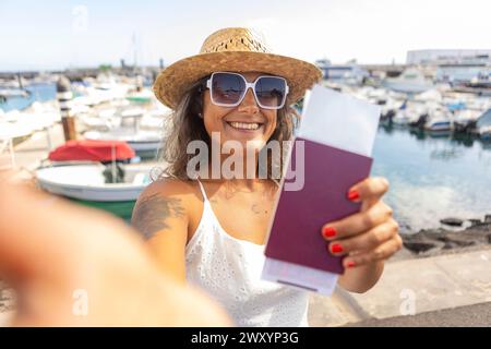 Fröhliche Frau in Sonnenbrille und Strohhut mit Pass, bereit für einen Wochenendausflug, mit Yachthafen Hintergrund. Stockfoto