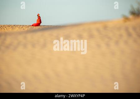 Eine einsame Frau in einem leuchtend roten Kleid sitzt anmutig auf einer Sanddüne in der Wüste und blickt in die Ferne. Stockfoto