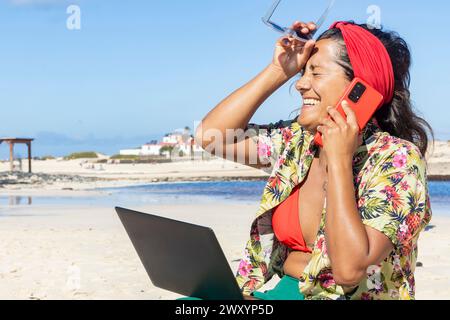 Ein fröhlicher digitaler Nomad genießt ein Telefongespräch, während er eine Sonnenbrille und einen Laptop an einem malerischen Strand hält Stockfoto