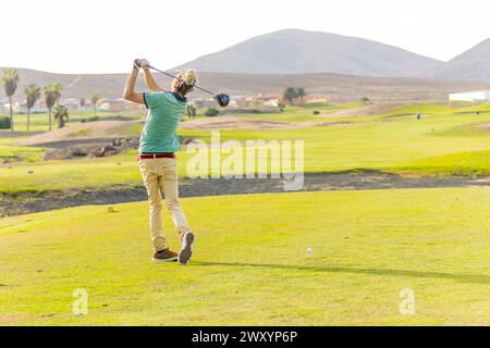 Die Rückansicht eines unverwechselbaren Golfers mit blonden Dreadlocks wird mitten im Schwung auf einem landschaftlich reizvollen Golfplatz erfasst und zeigt einen nicht-traditionellen Look im Sp Stockfoto