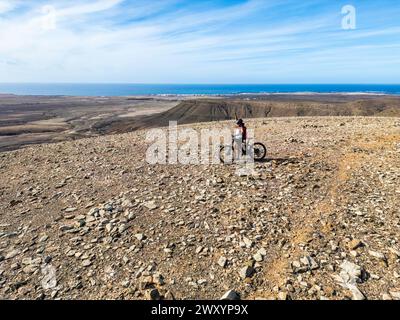 Eine Radfahrerin steht mit ihrem Mountainbike auf einem felsigen Bergweg und blickt unter klarem blauen Himmel auf das ferne Meer Stockfoto