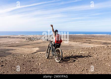 Ein Mountainbiker winkt von einem unbefestigten Weg mit einem Panoramablick auf die Küste im Hintergrund unter einem klaren blauen Himmel Stockfoto