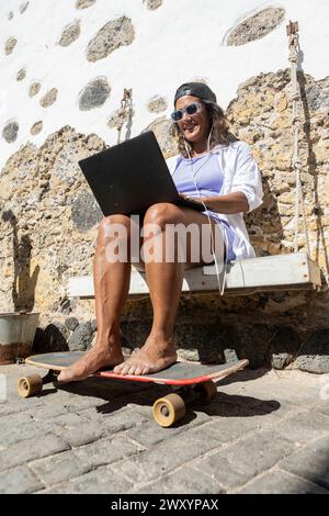 Von unten aus arbeitet eine lächelnde Frau mit Sonnenbrille und einer Kappe an ihrem Laptop, während sie auf einer Schaukel sitzt, die an einer rustikalen Wand hängt, mit einem Skateboard in der h Stockfoto