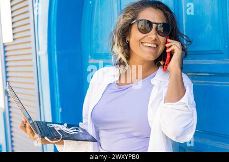 Eine lächelnde digitale Nomadin in legeren Kleidern arbeitet an ihrem Laptop, während sie am Telefon spricht und sich gegen eine blaue Wand lehnt. Stockfoto