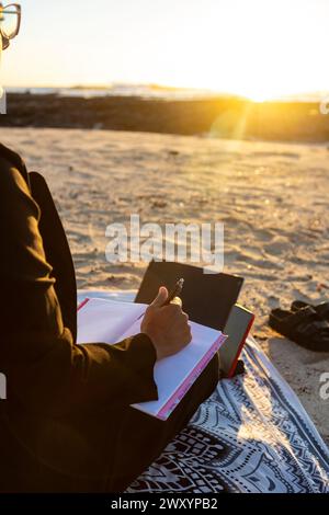 Der zugeschnittene, nicht erkennbare digitale Nomade arbeitet bei Sonnenuntergang auf einem Tablet an einem Sandstrand und verkörpert ein mobiles Büro. Stockfoto