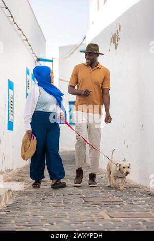 Multiethnische Paare machen einen gemütlichen Spaziergang mit ihrem liebenswerten Hund auf einer kopfsteingepflasterten Straße, umgeben von weißen Wänden und blauen Akzenten, die Sie präsentieren Stockfoto