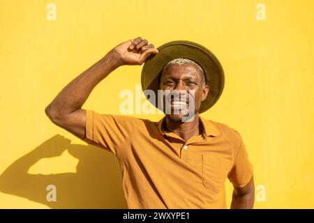 Ein fröhlicher Afroamerikaner mit einem hellen Lächeln, der seinen Hut hält, steht vor einem leuchtend gelben Hintergrund Stockfoto