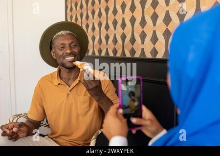 Ein fröhlicher Mann mit Hut verspeist ein Gebäck während er fotografiert wird und zeigt einen freudigen Moment, der mit der Kamera aufgenommen wurde Stockfoto