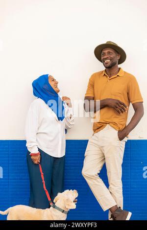 Ein fröhliches, multiethnisches Paar lacht zusammen, während es mit seinem Hund an einer kontrastierenden blau-weißen Wand steht Stockfoto