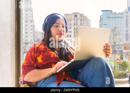 Eine junge Frau sitzt bequem am Fenster in einer städtischen Umgebung, arbeitet mit Kopfhörern an ihrem Laptop und blickt von der Kamera weg. Stockfoto