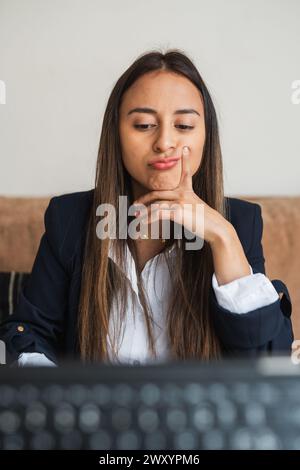 Eine junge Geschäftsfrau wirkt nachdenklich, während sie an ihrem Schreibtisch sitzt und ihr Kinn auf der Hand hält und in die Kamera blickt Stockfoto