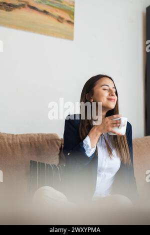Eine junge Frau in einem Business-Anzug genießt einen ruhigen Moment mit geschlossenen Augen und genießt ein heißes Getränk in einer gemütlichen Umgebung Stockfoto