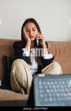 Eine junge Frau scheint desinteressiert zu sein, während sie am Telefon spricht, auf einer Couch in einem zwanglosen Zuhause sitzt Stockfoto