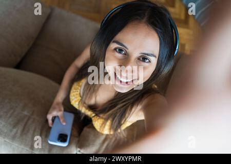 Die lächelnde junge Frau, die Kopfhörer trägt und ein Smartphone hält, macht ein Selfie, während sie zu Hause auf einer gemütlichen Couch sitzt Stockfoto