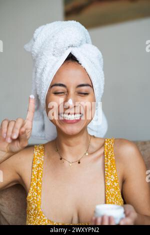 Eine fröhliche Frau mit einem Handtuch auf dem Kopf trägt Gesichtscreme auf und genießt ihre morgendliche Schönheitsroutine Stockfoto