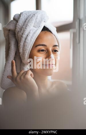 Eine junge Frau mit einem Handtuch auf dem Kopf gibt ihr Feuchtigkeit auf das Gesicht und sieht in der Nähe eines Fensters ruhig aus Stockfoto