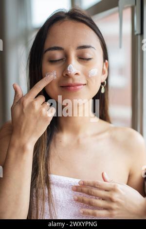 Eine junge Frau mit einem ruhigen Ausdruck trägt Gesichtscreme auf, die eine tägliche Schönheitsbehandlung hervorhebt Stockfoto