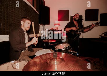 Zwei männliche Musiker treten in einer intimen Umgebung auf, einer spielt Schlagzeug und blickt weg, der andere singt und spielt Gitarre, vor der Kamera. Stockfoto