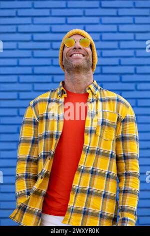 Ein lächelnder Mann in einem gelb karierten Hemd und einer roten Sonnenbrille genießt einen sonnigen Tag, der vor einer leuchtend blauen Ziegelmauer steht Stockfoto