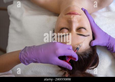 Schöne Frau erhält Schönheitsspritzen in die Stirn mit geschlossenen Augen. Der Kosmetikarzt händet Schönheitsoperationen an weiblichem Gesicht mit einer Spritze aus Stockfoto