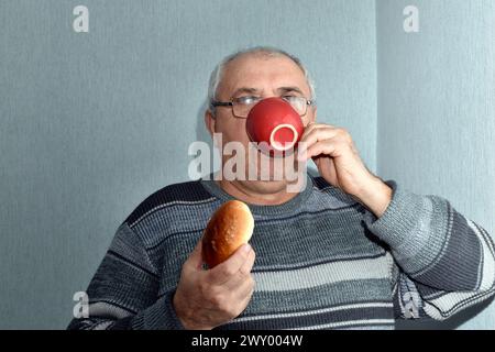 Das Bild zeigt einen älteren Mann, der aus einem Becher trinkt und ein Brötchen in der Hand hält. Stockfoto