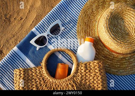 Sommeraccessoires am Sandstrand bei Sonnenuntergang. Gewebte Tasche mit Sonnencreme innen neben einem stylischen Sonnenhut. Weiße Sonnenbrille liegt auf einem blau gestreiften Handtuch Stockfoto