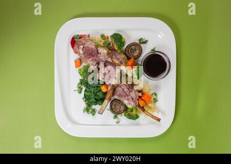 Blick von oben auf ein Gericht mit saftigen Lammrippchen, im Ofen gebackenem Gemüse und duftender roter Sauce auf grünem Hintergrund Stockfoto