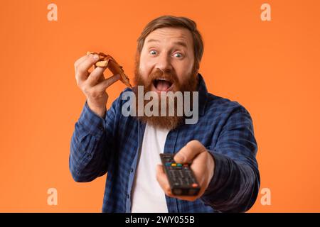 Rothaariger bärtiger Kerl zeigt Fernbedienung, während er Pizza und Studio genießt Stockfoto