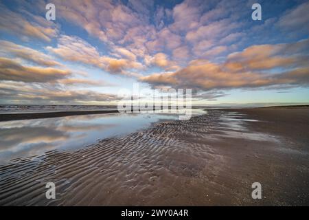 Spektakuläres Bild der Küste unter einem Wolkenhimmel, der das rote Leuchten der Sonne zeigt. Am teilweise trockenen Strand sind Wasserpfützen zu sehen Stockfoto