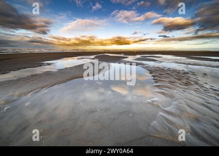 Spektakuläres Bild der Küste unter einem Wolkenhimmel, der das rote Leuchten der Sonne zeigt. Am teilweise trockenen Strand sind Wasserpfützen zu sehen Stockfoto