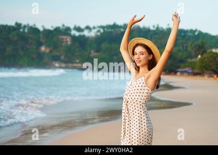 Glückliche Frau mit Strohhut, Polka Dot Kleid hebt Arme am Sandstrand. Lächelnde Frau genießt tropische Sonne, Wellen, Urlaub am Meer. Alleinreisende Stockfoto