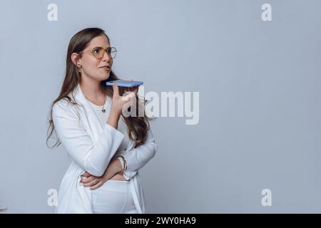 Junge lateinfrau in weißer Kleidung, mit langen Haaren und Brillen, glückliche Geschäftsfrau, die steht und eine Sprachnachricht mit ihrem Telefon sendet, Studio-Sho Stockfoto