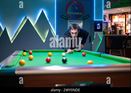 Ein junger Mann in schwarz gekleidet und lächelnd, der in einer dunklen Cocktailbar Billard spielt. Es ist eine warme Bar mit Neonlichtern. Stockfoto