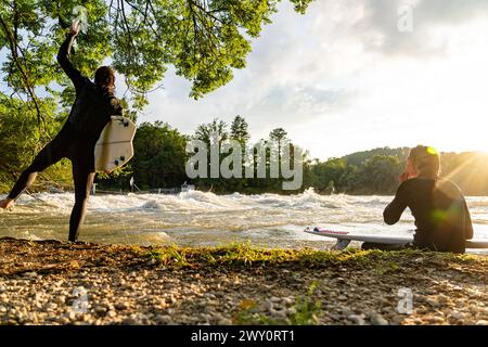 Ein junger Surfer am Fluss Aare genießt die Ruhe des Sonnenuntergangs, während ein anderer sein Brett auf Felsen balanciert. Stockfoto