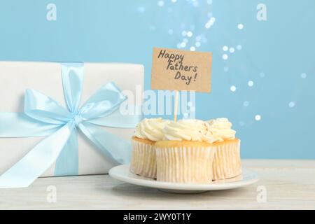 Alles Gute zum Vatertag. Leckere Cupcakes mit Grüßen und Geschenkbox auf hellem Holztisch, Nahaufnahme Stockfoto